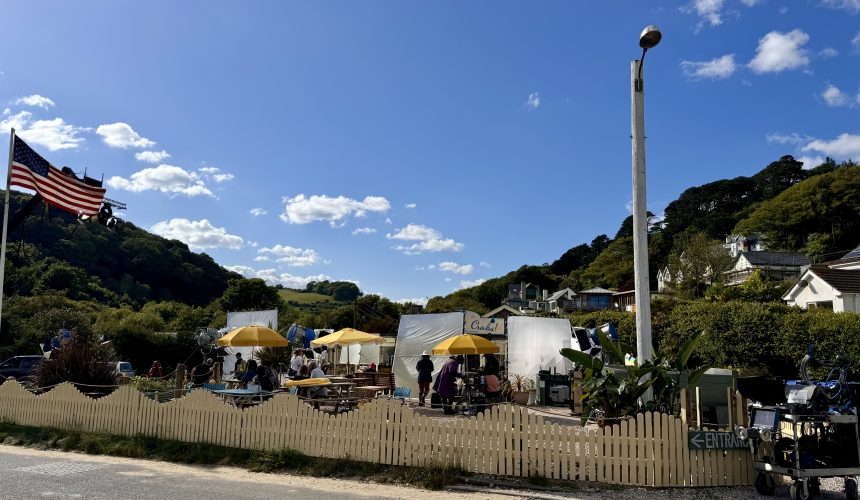 Bright outdoor scene at a festival with a stage, yellow umbrellas, and scenic hillside houses in the background.